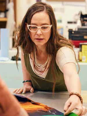 A woman wearing glasses and a necklace examines a colorful print on a table in an art studio, surrounded by art supplies.